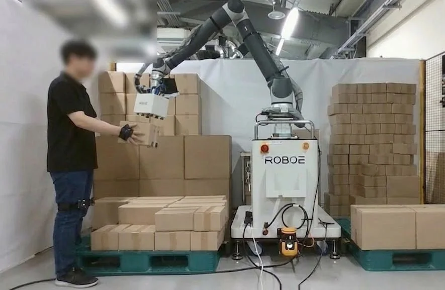A worker in motion-capture gloves reaching for a box while a palletizing robot works in the same space